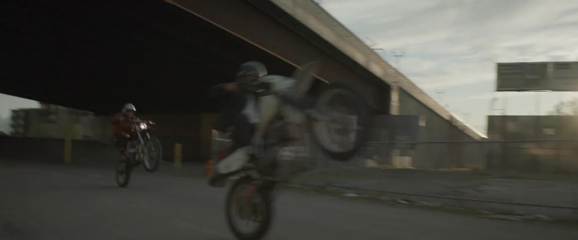 Two motorcyclists pop wheelies while riding under a freeway pass