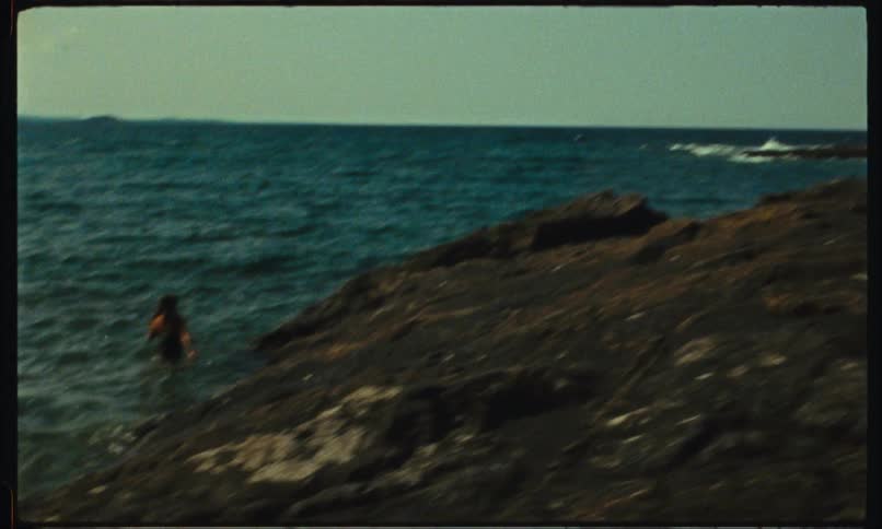 A little girl points to a person who is in the sea water next to rocks