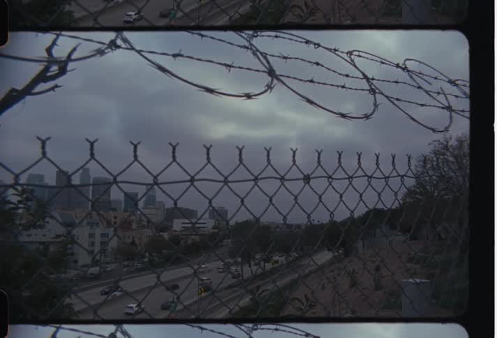 A gloomy scene of a city and a highway seen through a fence with barbed wire on top