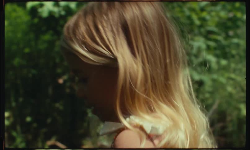 A girl with blonde hair is outdoors surrounded by greenery and sunlight
