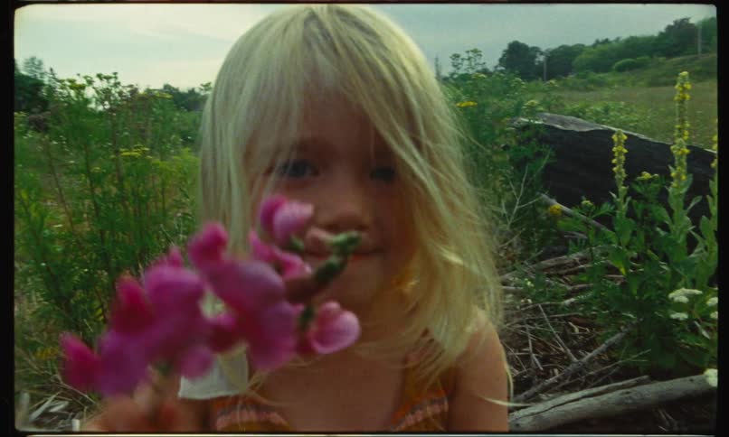 A little girl stands in a field and smiles as she smells pink flowers