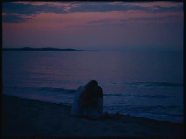 Mother embracing her daughter sitting by the sea with a reddish sky in the background
