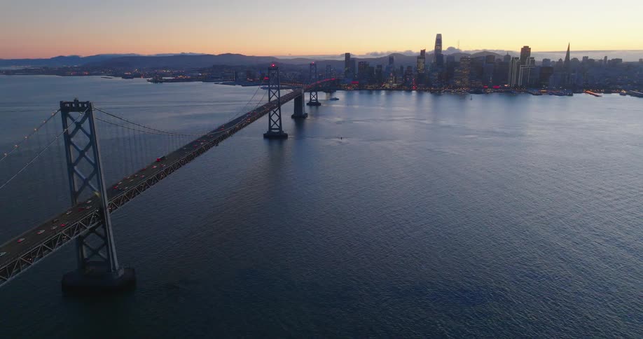 Traffic crosses suspension bridge over ocean toward downtown skyline under a red tinted horizon