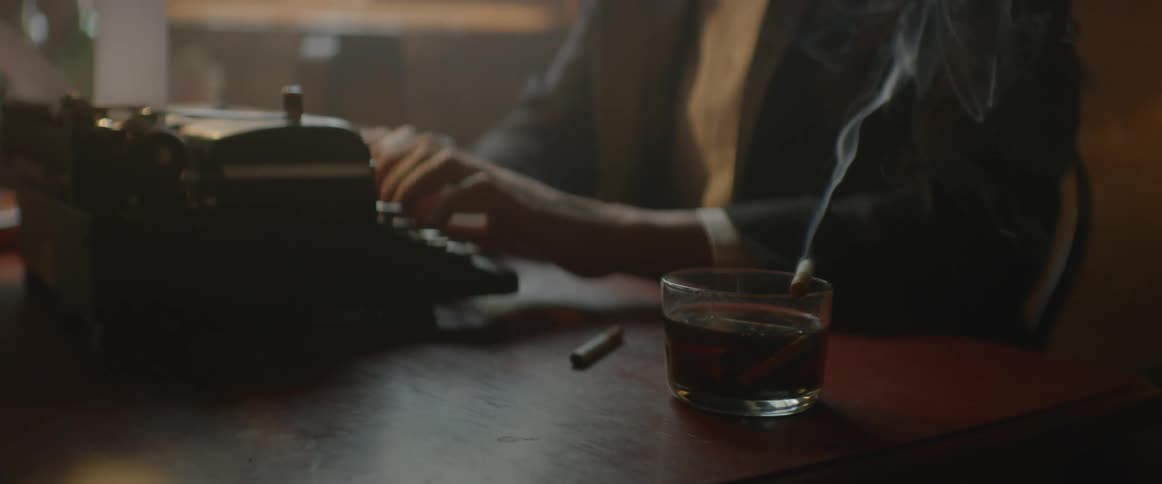 A man types at a typewriter as a cigarette burns on the desktop