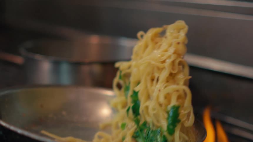 Chef holds skillet handle with towel while flipping pasta cooking in skillet over fire
