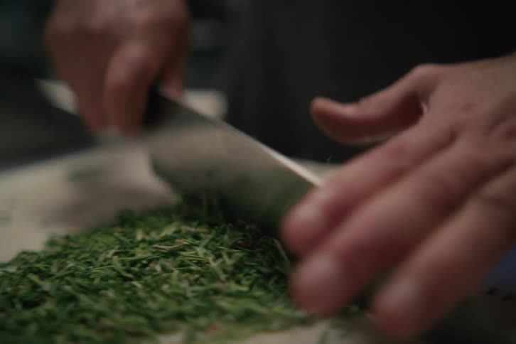 A pair of hands using a large kitchen knife to chop herbs for seasoning