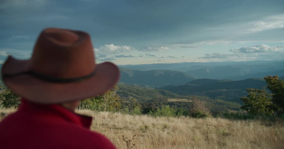 Older cowboy stands in field overlooking foggy mountainous terrain on a cloudy day