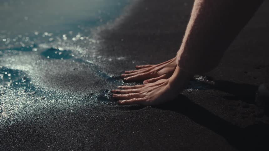 A woman bent down on a beach grasping at the sand at the edge of the sea