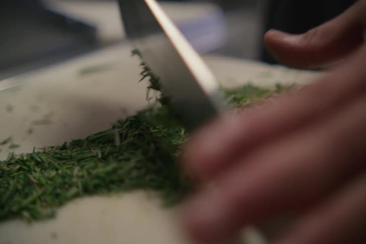 A pair of hands using a large kitchen knife to chop herbs for seasoning