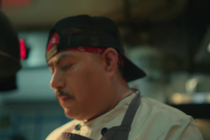 A man with a backwards hat and mustache is looking down cooking something