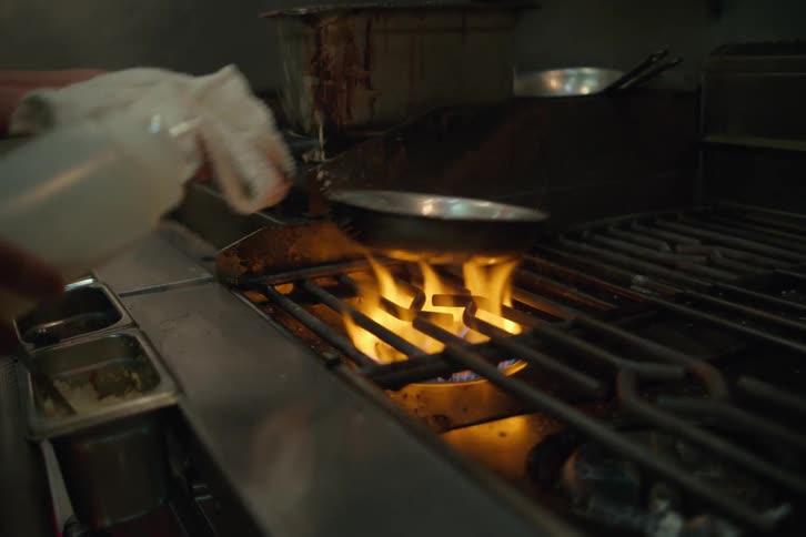 A person is pouring liquid into a pan that is frying something and there is a high flame under the pan