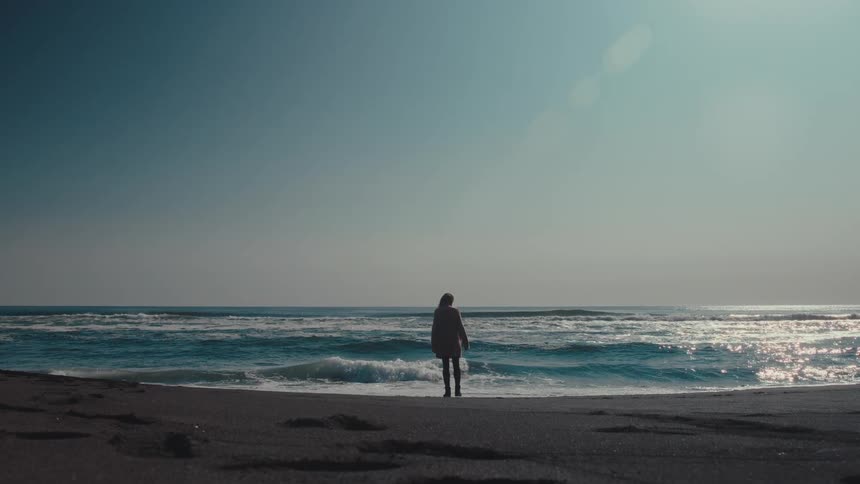 A woman standing on the beach looking at the ocean waves in a serene scene