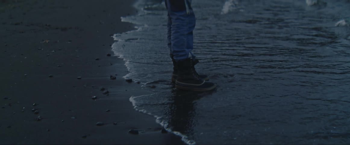 A person in a yellow jacket walking along the seashore until a small wave reaches their feet