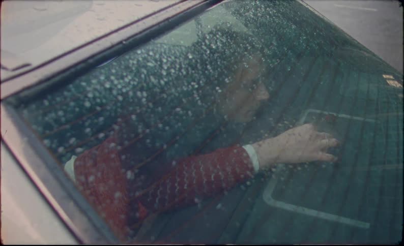 A woman looks out the back window of the car as it rains