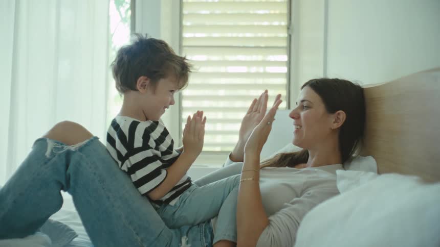 Mother and son playing indoors on bed clapping each others hands in a playful manner during day time