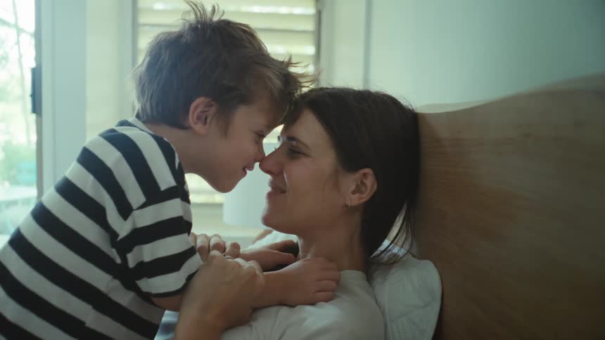 Mother lying on bed rubs noses with her son as they smile