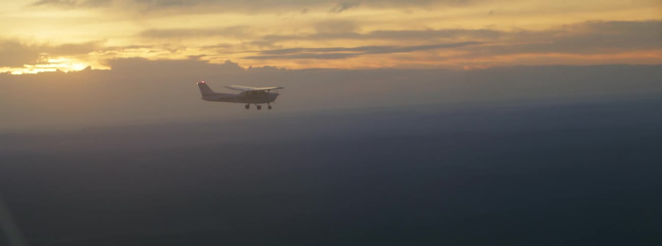 A small plane flying over the land with a cloudy sky at sunset