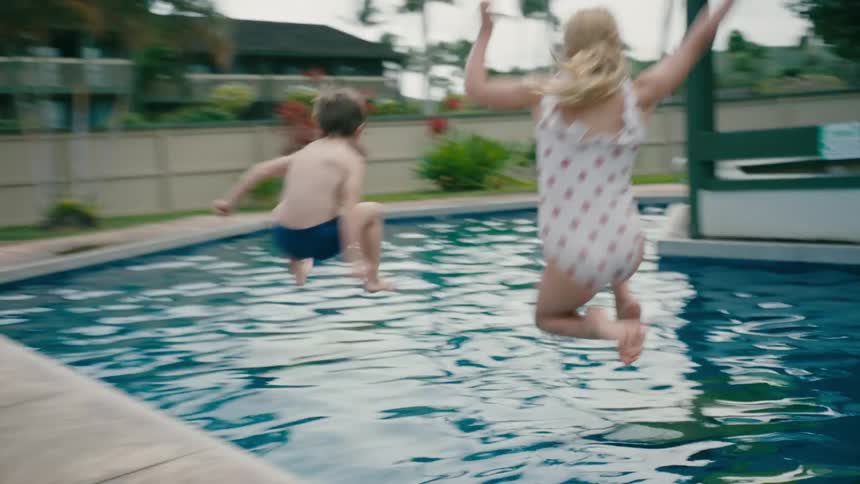 Kids jump into backyard swimming pool as parents sit nearby