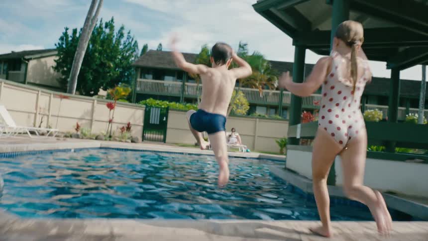 A little boy and girl run side by side over grass and patio to jump splashing into a hotel pool
