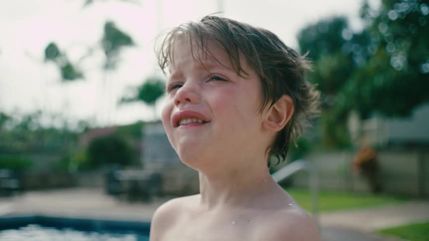 Boy standing outdoors next to a pool on a lawn crying during day time