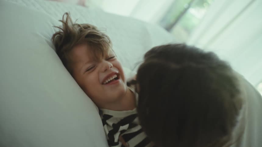 A mother rolls her son onto a bed and kisses him playfully