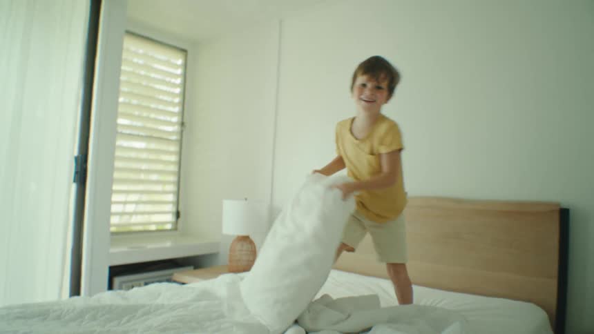 Boy standing on the bed pillow fights with the camera