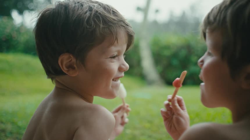 Two children licking ice cream while seated on the grass outdoor during the day