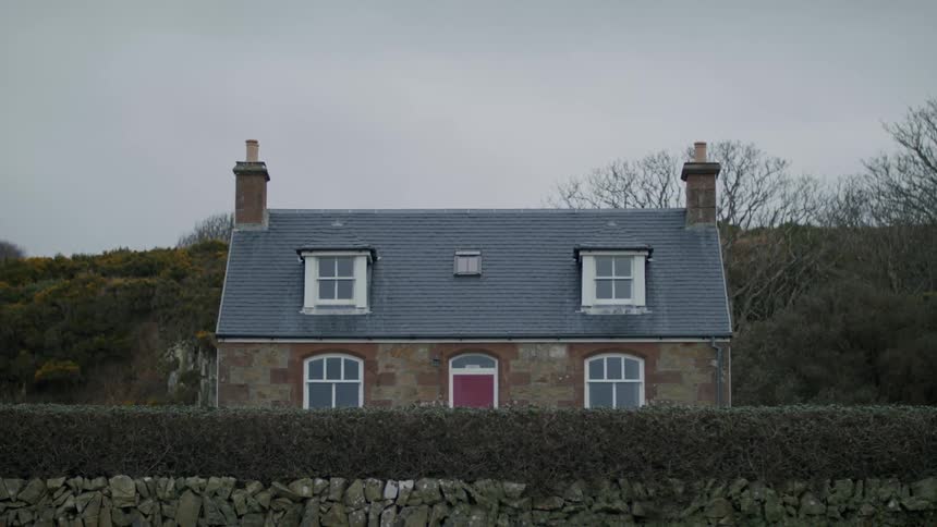 Hunting lodge home with smoke stacks on both sides and a red door on a hillside behind a stone wall on a cloudy day