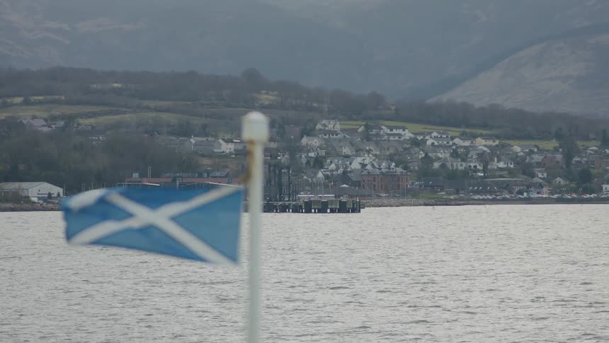 A blue flag with a white cross on a pole near a calm lake and a coastal town