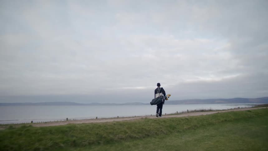 Man carrying a golf bag walks on a road next to a lake