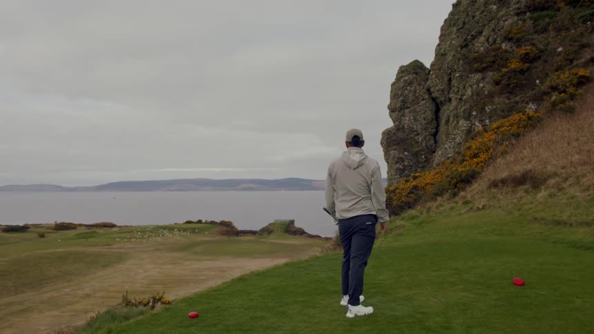 A golfer on a grassy field near a large rock formation and a calm body of water