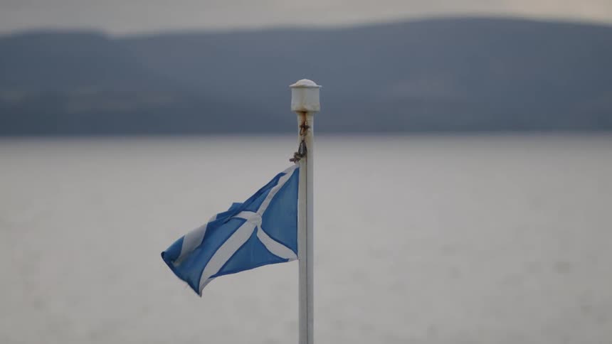 A blue flag with a white cross on a pole near a calm lake and distant mountains