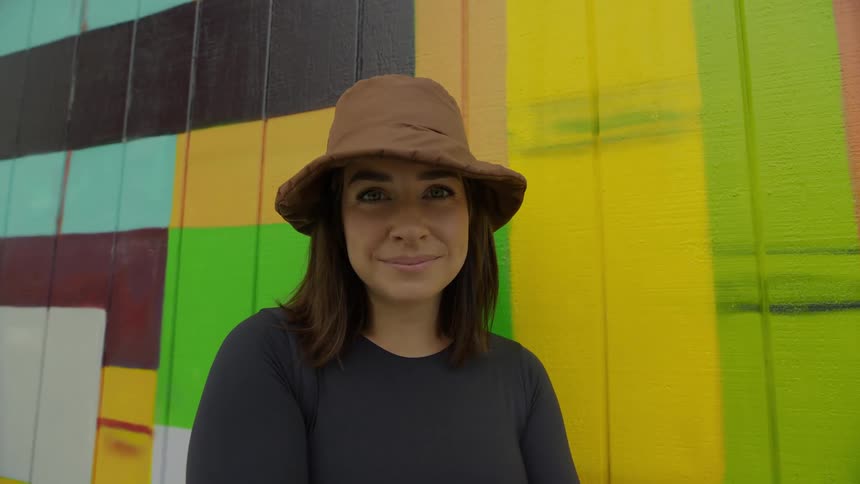 A woman in a hat stands and poses in front of a wall painted with art