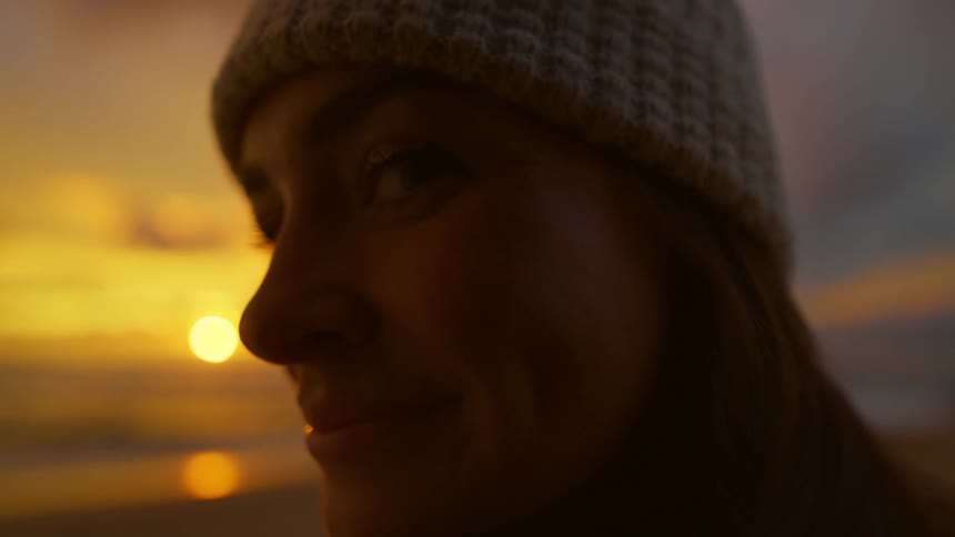 Woman smiles and looks to the side away from the sun setting over the ocean in the background