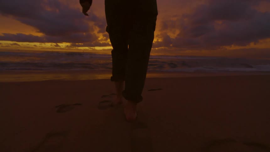 Silhouette of a person walking across the beach toward the water with their pants rolled as the sun has set