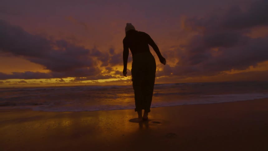 Silhouette of a teenage girl walking across the beach with her pants rolled up as the waves roll in and over her bare feet