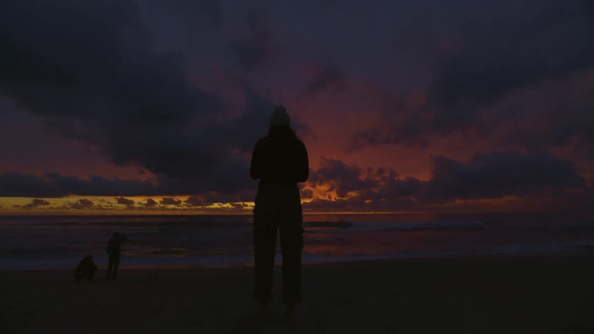 A woman stands on a beach and watches the sun setting over the ocean