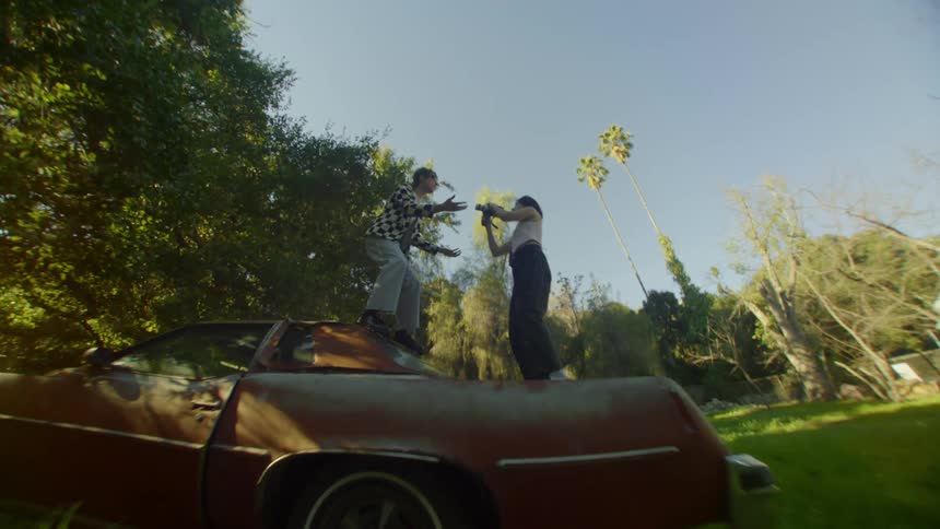 Woman records video of man singing while they stand on old rusted car in field
