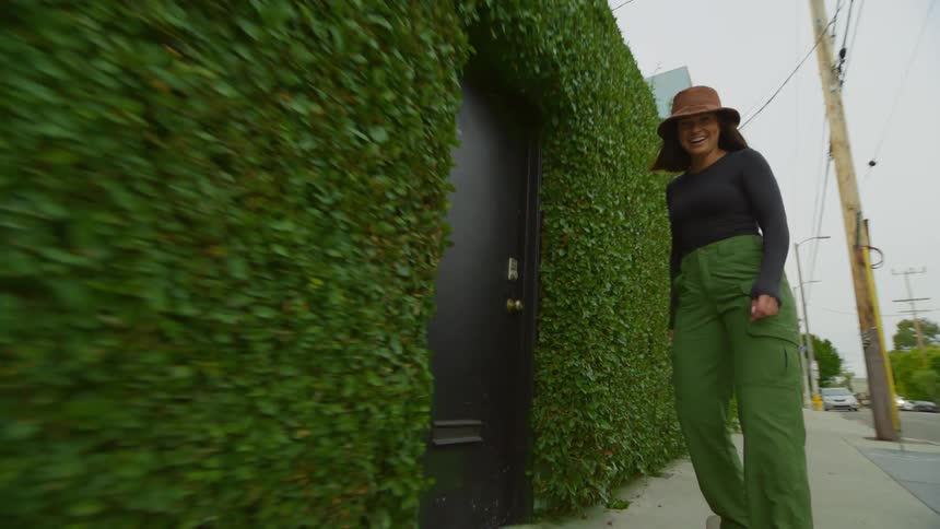 A woman skateboarding on a sidewalk next to a hedge and a parked van
