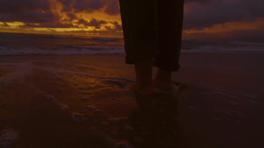 A person stands on a beach with their feet in the water at sunset