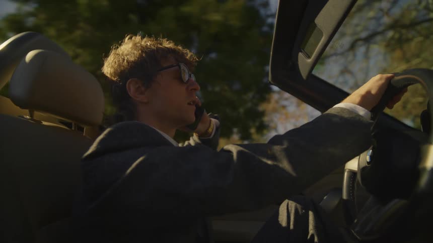 A man wearing sunglasses talks on his cell phone as he drives his convertible car