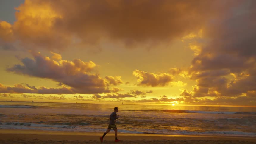 A jogger runs across the beach as boats sail and the sun glows on the horizon under a colorful sky