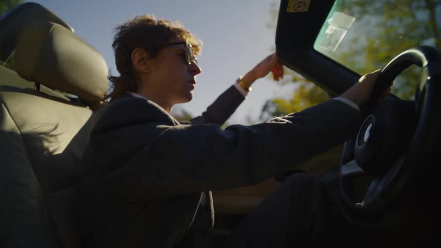 A man wearing sunglasses is driving a convertible car with one hand above the windshield and the other hand on the steering wheel