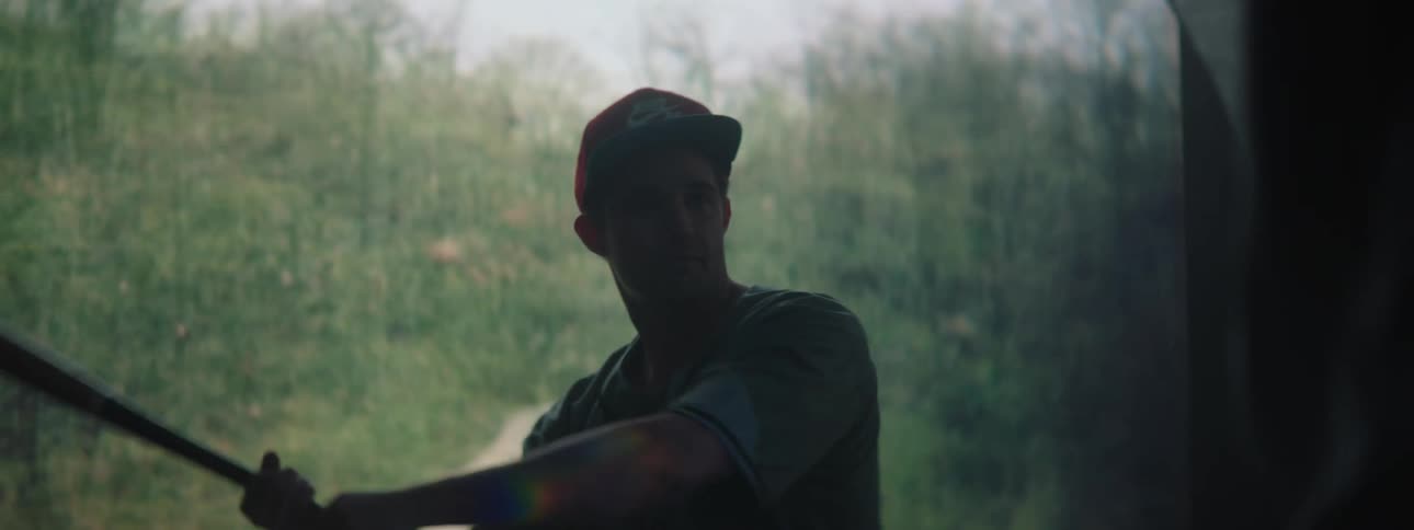 A man in a baseball uniform swings a bat slowly in front of a screen with a nature image on it