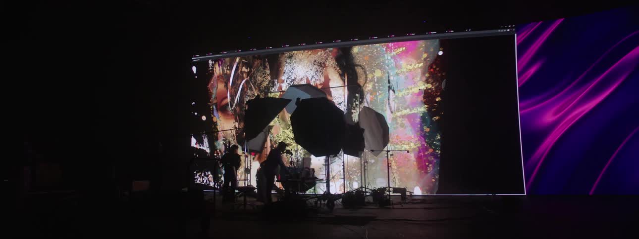 A pair of people set up a film set in front of a screen displaying artistic graphics
