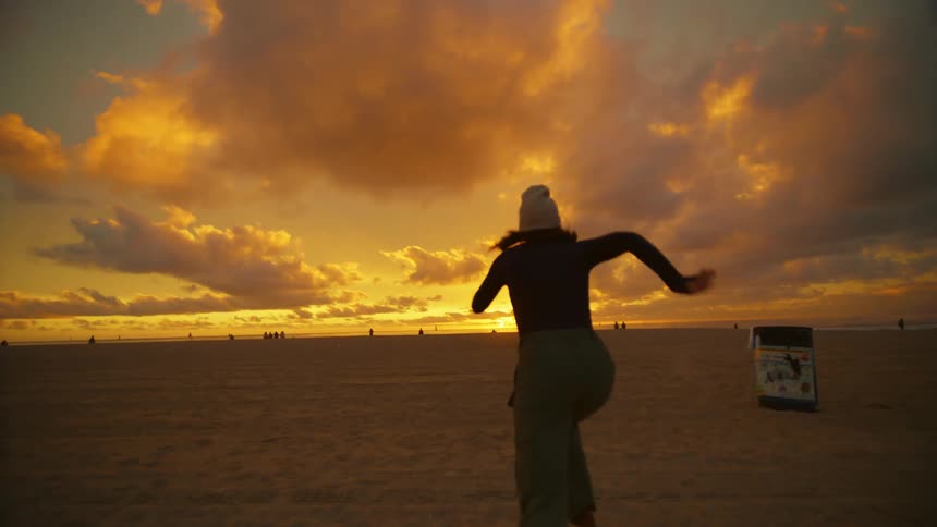 Young woman wearing a beanie hat runs towards the sunset at the beach