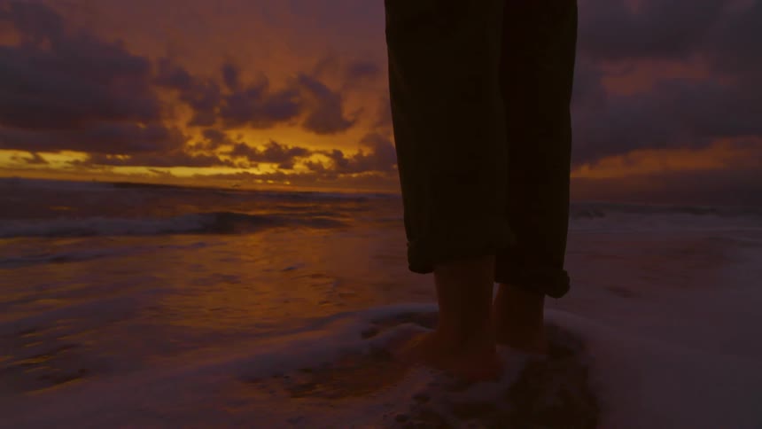 A person standing barefoot with pant legs rolled up on the beach at sunset getting their feet wet