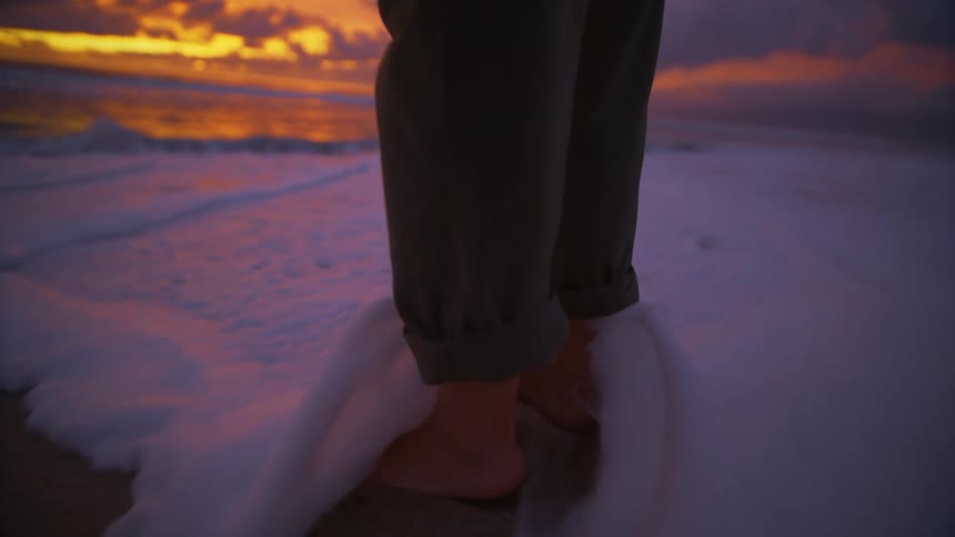 Bare feet of a person standing on the beach as waves roll into shore and over their feet as the sun has set on the horizon