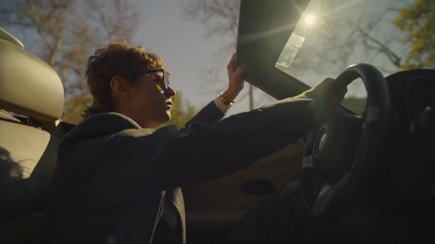 A man wearing sunglasses is driving a convertible car with one hand above the windshield and the other hand on the steering wheel