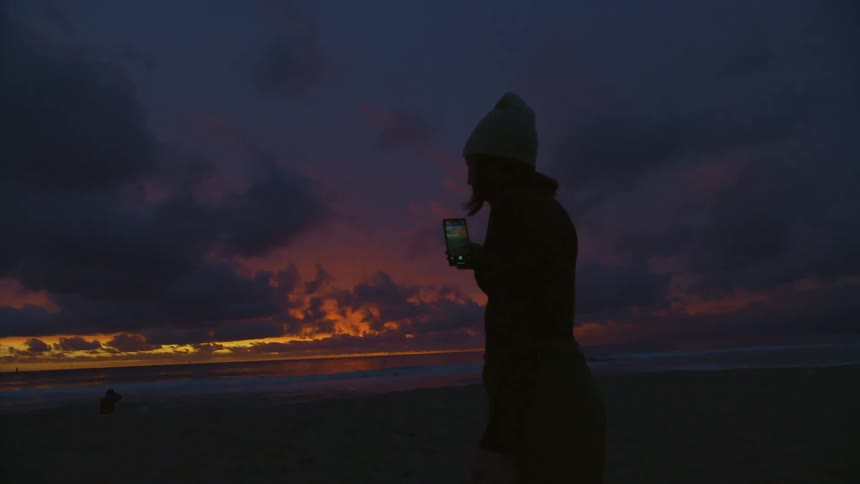 Silhouette of a teenage girl standing on the beach taking a picture with her phone as the sun has set
